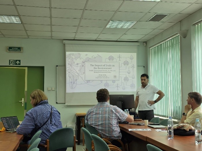 A presentation on "The Impact of Trade on the Environment" is taking place in a conference room with attendees seated at tables.