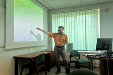 A person is giving a presentation in a room with a projector screen, pointing at a graph. There are chairs and a computer in the background.