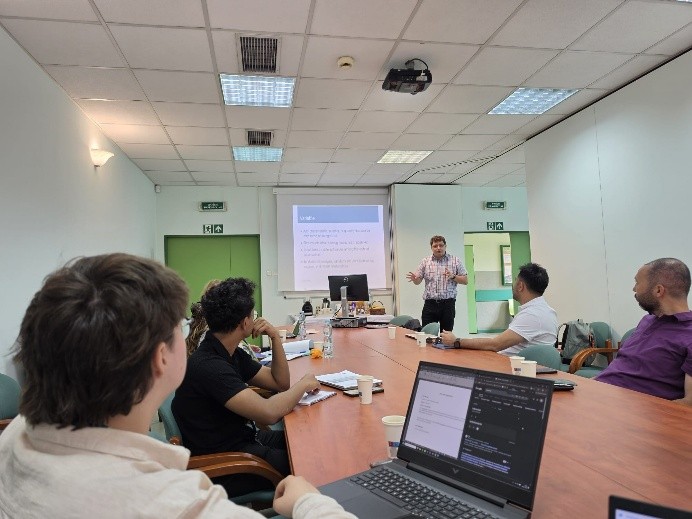 A group of people in a meeting room listens to a presenter discussing a topic, with a projector screen displaying information.