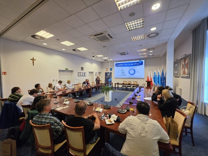 A meeting room with a long table, attendees seated, a presentation screen, and flags in the background.